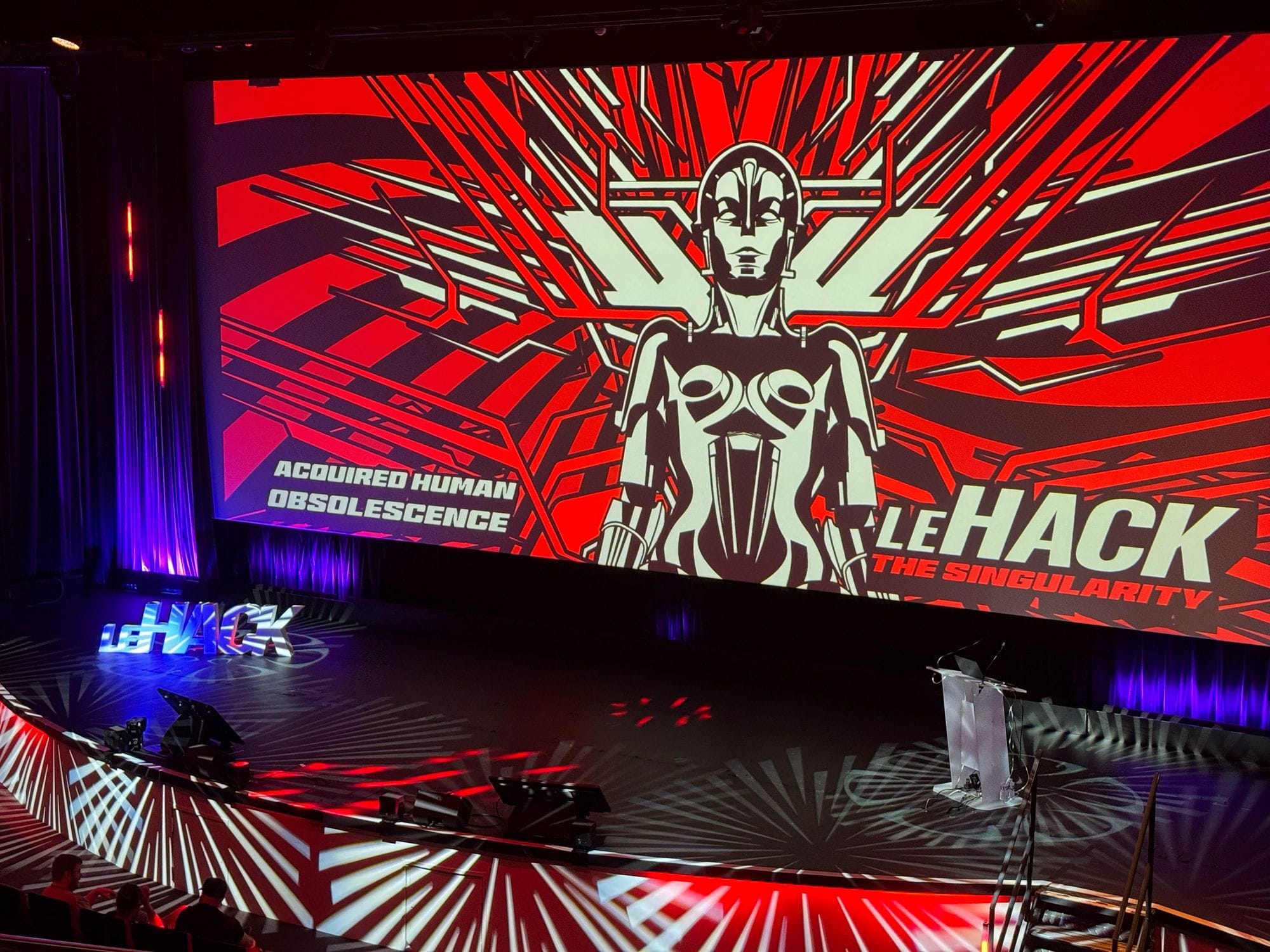 Photo de la scène où une image dit : "Acquired Human Obsolescence. LeHack. The Singularity."